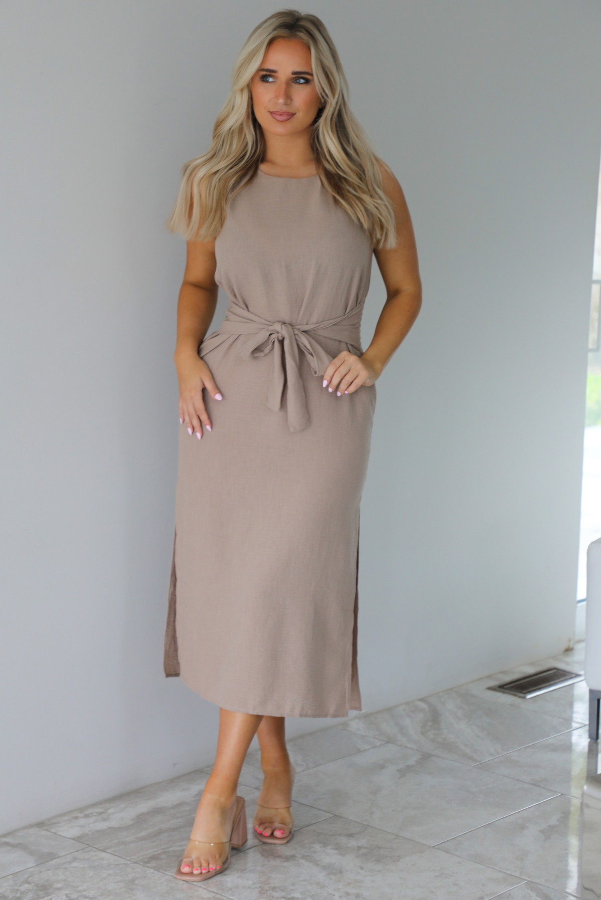 Woman wearing a tan, high neckline, midi dress that has a tie detail at the waistline and split hem details on either side. The photo is taken indoors against a white wall with white marble flooring. 