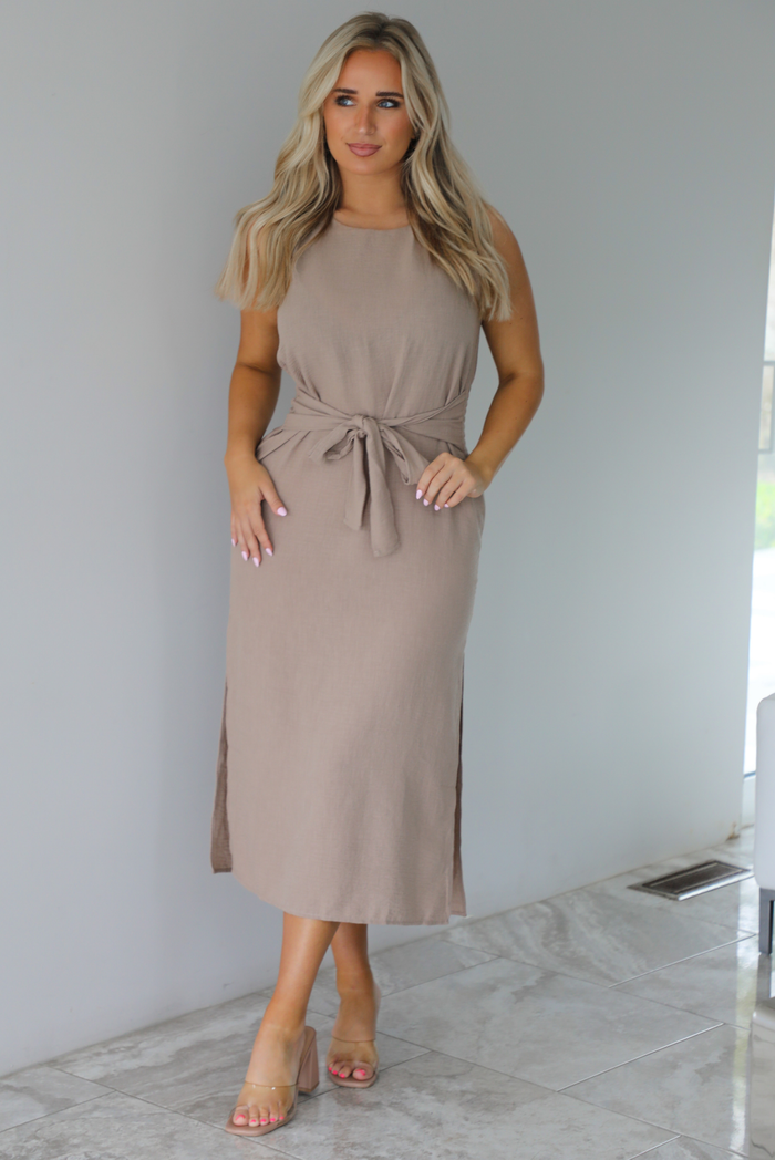 Woman wearing a tan, high neckline, midi dress that has a tie detail at the waistline and split hem details on either side. The photo is taken indoors against a white wall with white marble flooring. 