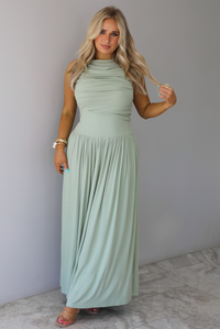 Woman wearing a sage green maxi length dress, which has a rouched detail at the bust, a fitted waistline, a flowy skirt. The photo is taken indoors against a white wall with white marble flooring. 