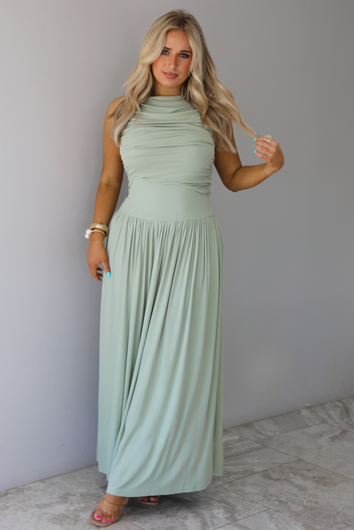 Woman wearing a sage green maxi length dress, which has a rouched detail at the bust, a fitted waistline, a flowy skirt. The photo is taken indoors against a white wall with white marble flooring. 