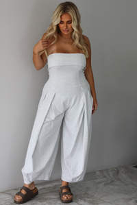 A woman modeling a light gray strapless jumpsuit with wide, balloon-style legs and gathered detailing, standing indoors against a white wall with white marble flooring. 