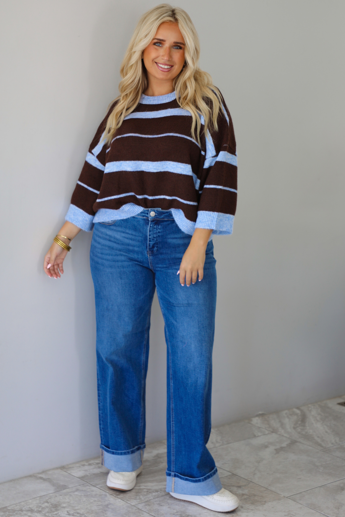 A woman wearing a brown and blue striped knit top with dark denim jeans, standing indoors against a white wall with white marble flooring. 