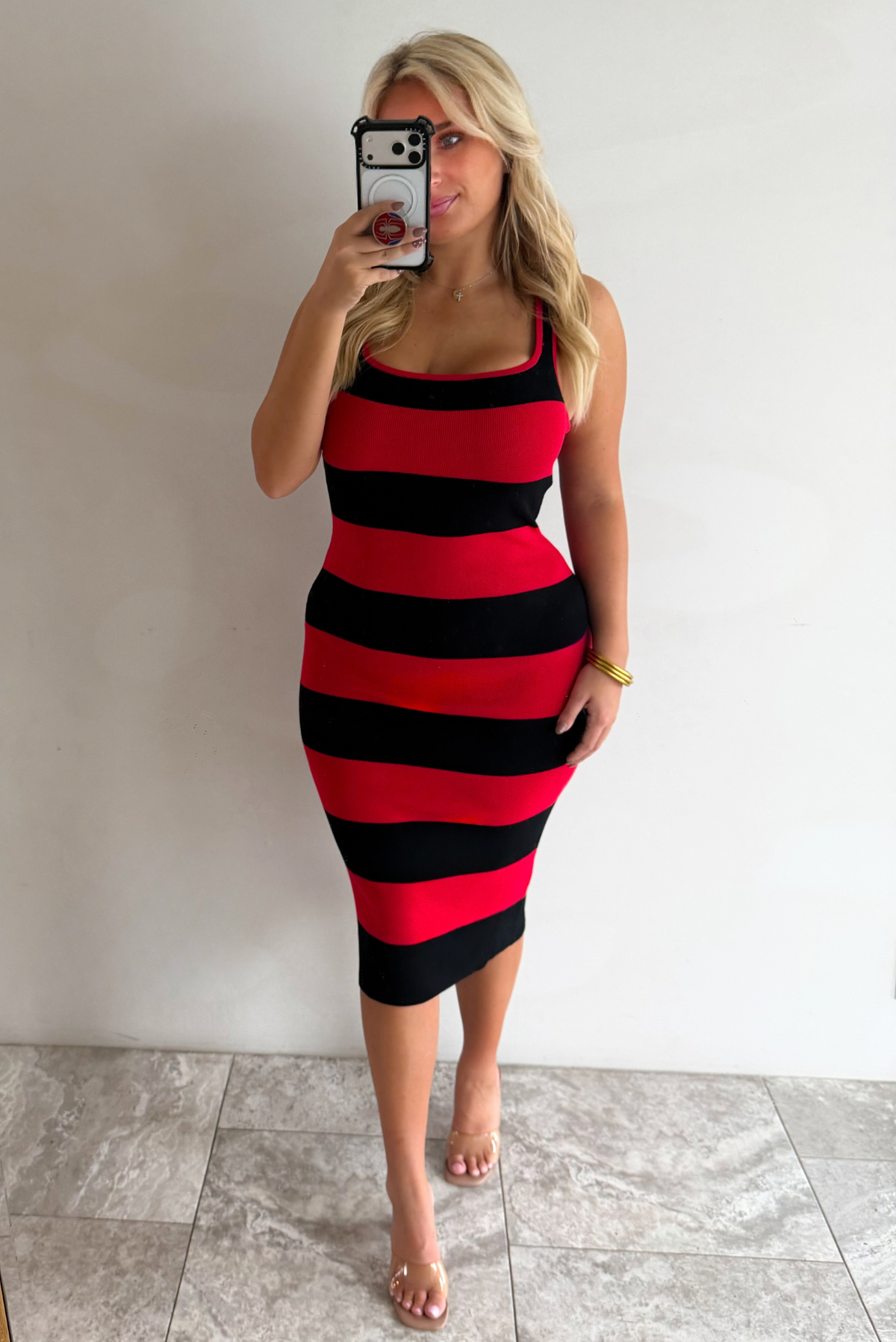 A woman taking a mirror selfie wearing a red and black striped body-con midi dress, paired with clear strapped heels, standing indoors against a white wall with white marble flooring. 