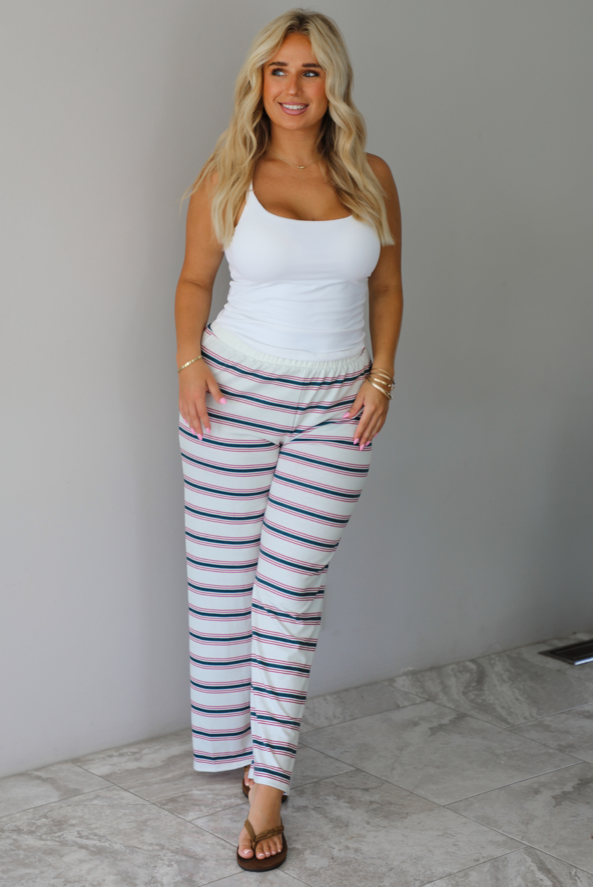 A woman modeling a pair of cream-white wide-leg pants with thin green and red horizontal stripes, paired with a white tank top and brown sandals, standing indoors against a white wall with white marble flooring. 