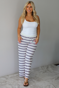 A woman modeling a pair of cream-white wide-leg pants with thin green and red horizontal stripes, paired with a white tank top and brown sandals, standing indoors against a white wall with white marble flooring. 