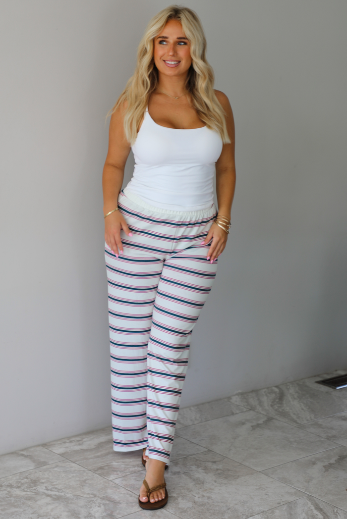 A woman modeling a pair of cream-white wide-leg pants with thin green and red horizontal stripes, paired with a white tank top and brown sandals, standing indoors against a white wall with white marble flooring. 