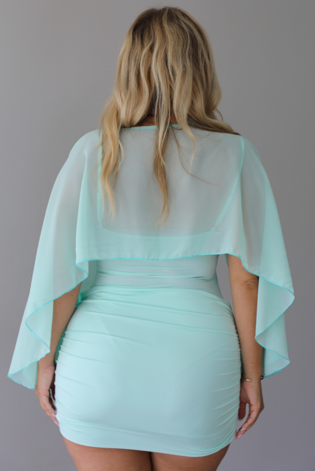 A woman shown from behind in a light mint green mini dress with a sheer draped shawl, standing indoor against a white wall.