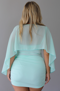 A woman shown from behind in a light mint green mini dress with a sheer draped shawl, standing indoor against a white wall.