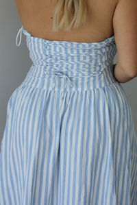 Woman wearing a blue and white striped strapless maxi dress indoors against a white wall. The photo shows a closeup of the corset-like lace up detailing in the back.