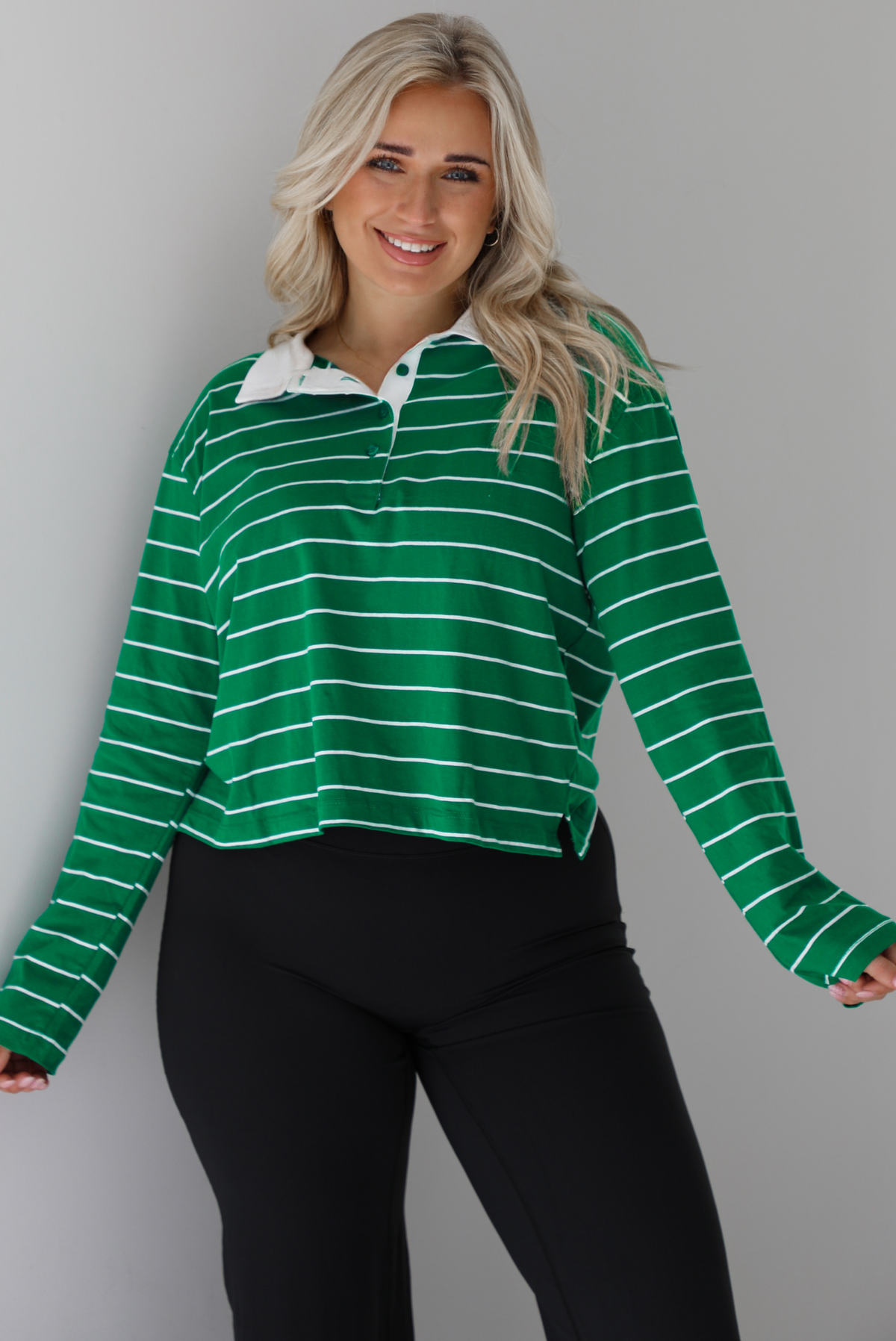 Woman wearing a green with white striped collared cropped top with functional button closures a quarter at the top. The top is paired with black leggings. The photo is taken in front of a white wall.