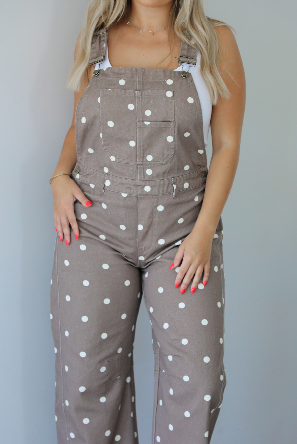 Woman wearing a brown with white polka dots overalls jumpsuit indoors against a white wall.