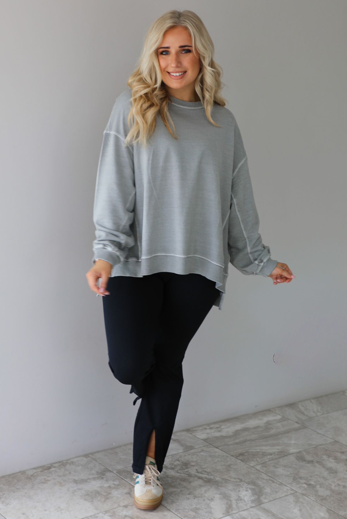 Woman wearing a grey oversized sweater with black leggings that have a slightly flared hem with split hem details in the front. The photo is taken in front of a white wall with white marble flooring. 