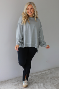 Woman wearing a grey oversized sweater with black leggings that have a slightly flared hem with split hem details in the front. The photo is taken in front of a white wall with white marble flooring. 