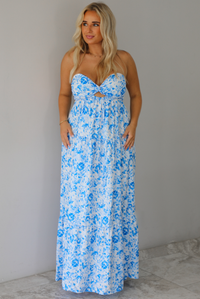 A woman modeling a strapless white maxi dress with a blue floral print and a sweetheart neckline with a small keyhole cutout, standing indoors against a white wall with white marble flooring. 