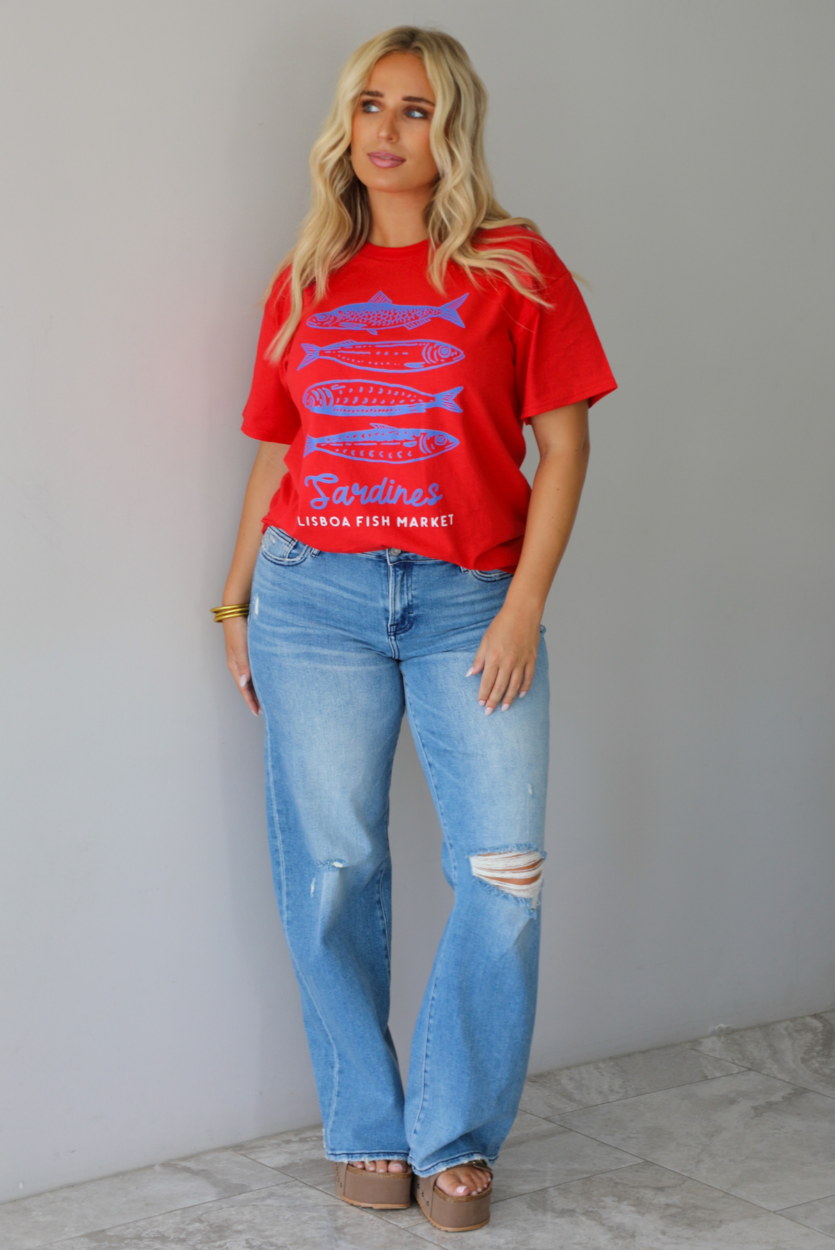 A woman wearing a red graphic t-shirt with blue sardine illustrations and the text 'Sardines LISBON FISH MARKET', paired with blue jeans, standing indoors against a white wall with white marble flooring.