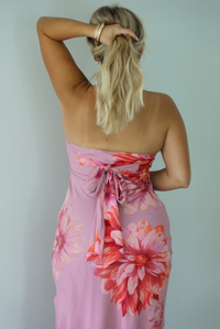 A woman viewed from behind wearing a strapless mauve maxi dress with a large red and orange floral print and a tie detail at the back waist, standing indoors against a white wall.