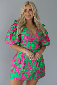 Woman wearing a fuchsia and green dress that has puffed sleeves, a v-neckline, fitted bodice, and flared skirt with pleated details indoors against a white wall.