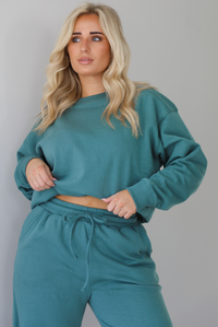 Woman wearing a sage two piece sweat-set. The set features a slightly oversized sweater and high waisted sweat pants that have an elastic and drawstring waistline with pockets. The sweater is tucked in. The photo is taken indoors against a white wall with white marble flooring. 