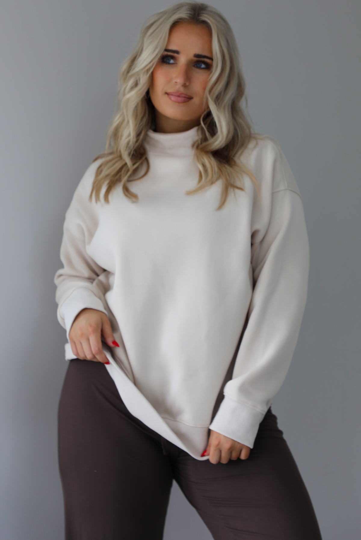 Woman wearing a beige high-neck, oversized sweaters and high waisted brown pants. The photo is taken indoors against a white wall with white marbled flooring. The photo shows a closeup of the oversized sweater untucked.