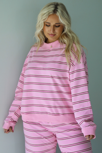 A woman wearing a pink two piece sweat set featuring a maroon and white striped design on a sweatshirt and high waisted, wide leg pants, standing indoors against a white wall. The photo shows a closeup of the sweatshirt, which has a thick ribbed neckline and the same thick ribbed material at the wrists and bottom hem. 