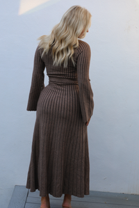 Woman wearing a taupe colored dress that has a vertical ribbed pattern throughout, a high neckline, fitted bodice, cinched waistline, long sleeves that flare slightly, and a maxi length skirt that flows at the bottom. The photo is taken outdoors against a white wall.  The photo shows the back of the dress, which looks like the front. 