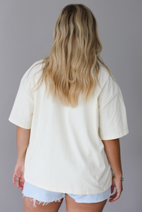 Woman wearing a graphic t-shirt with a lobster wearing a cowboy hat and the words 'HOWDY CLAW' written along the front and denim shorts against a white wall. The photo shows the back of the tee. 