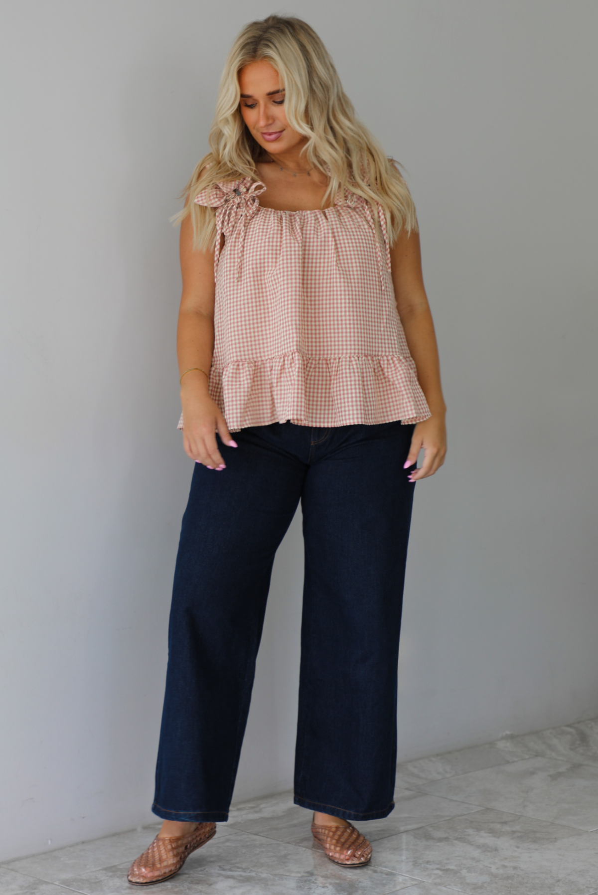 A woman wearing a light maroon and cream gingham flowy top with dark denim jeans, standing indoors against a white wall with white marble flooring.