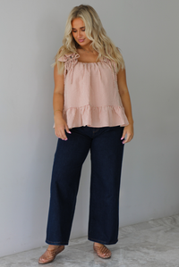 A woman wearing a light maroon and cream gingham flowy top with dark denim jeans, standing indoors against a white wall with white marble flooring.