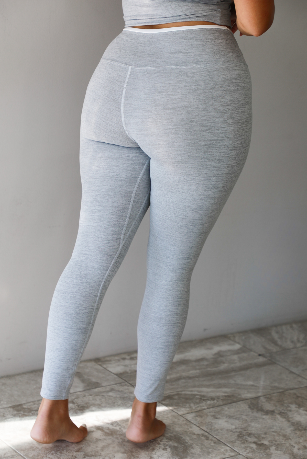 A woman modeling a light gray two-piece loungewear set consisting of a cropped tank top and matching leggings, focused on a closeup of the back of the leggings, standing indoors against a white wall with white marble flooring. 