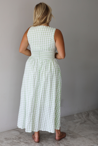 A woman shown from behind wearing a light green and white gingham maxi dress with a drop waist bodice and side pockets, standing indoors against a white wall with white marble flooring. 