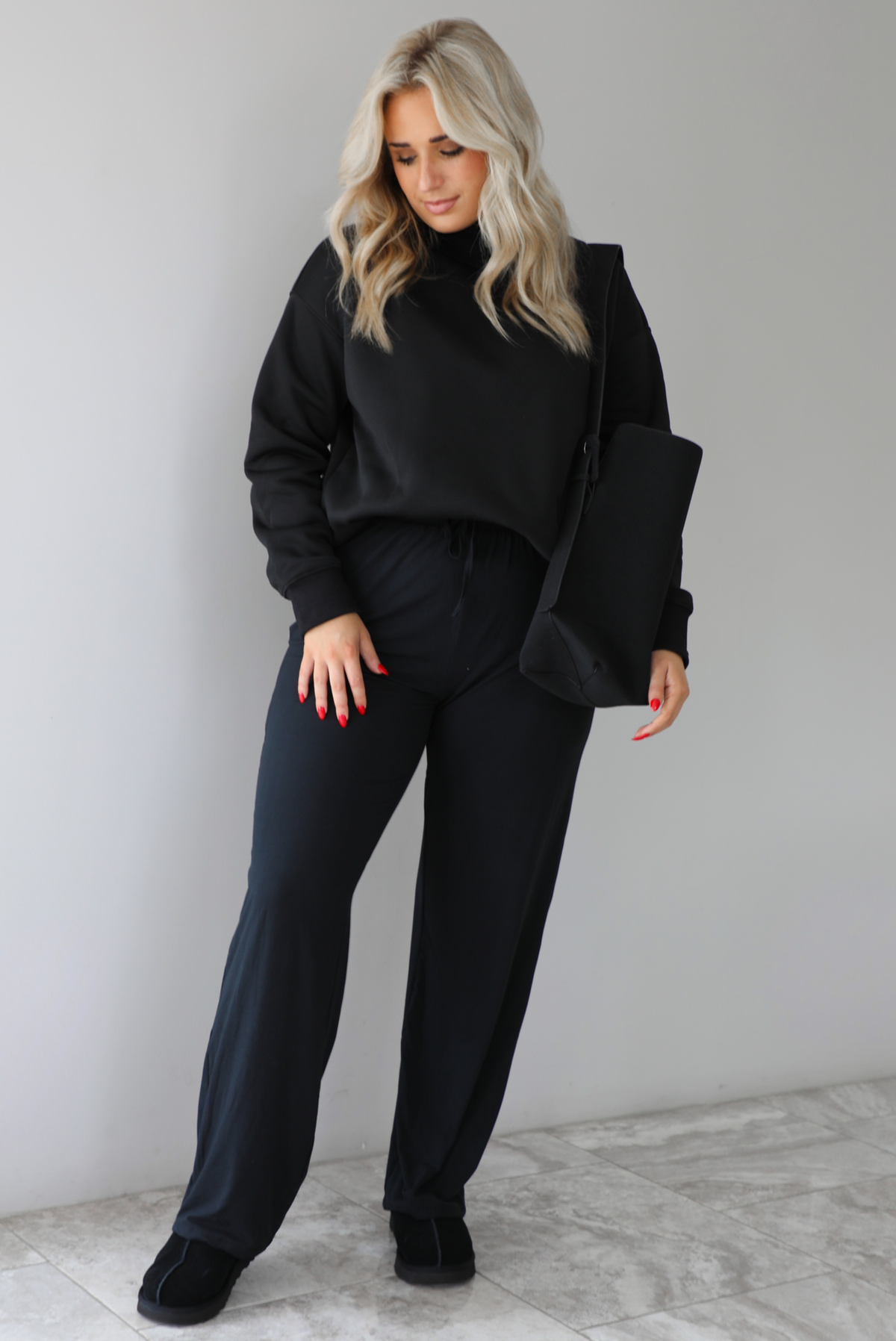 Woman wearing a black high-neck, oversized sweater, high waisted black pants, with a black shoulder bag, and black ugg boot shoes. The photo is taken indoors against a white wall with white marbled flooring. 