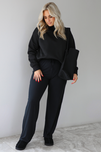 Woman wearing a black high-neck, oversized sweater, high waisted black pants, with a black shoulder bag, and black ugg boot shoes. The photo is taken indoors against a white wall with white marbled flooring. 
