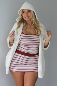 A woman wearing a cream colored oversized cardigan worn over a striped two piece set, standing indoors against a white wall. The photo shows the model wearing the hoodie detail on the cardigan. 