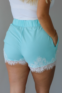 A person wearing light aqua blue shorts with white lace trim at the hem, shown from the back against a white wall. 