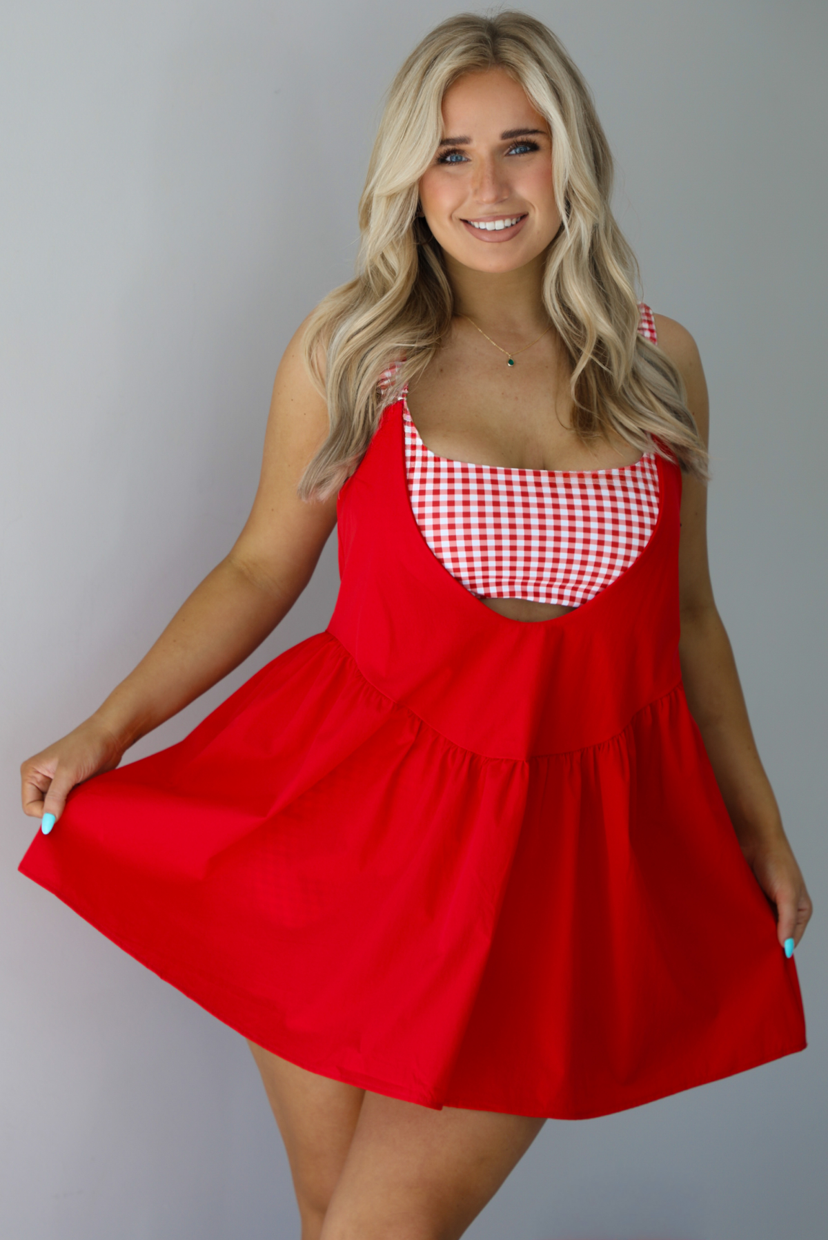 Woman wearing a red dress with a red and white gingham romper underneath. The photo is taken indoors against a white wall.