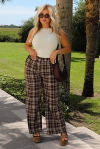 A woman stands outdoors on a paved walkway wearing a sleeveless cream top, brown, cream, and sand plaid wide-leg trousers, sunglasses, and brown platform sandals, with a dark brown shoulder bag.
