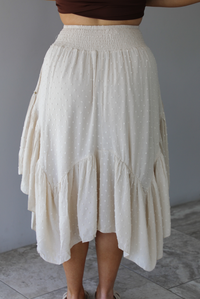 Woman wearing a sand colored midi skirt featuring a smocked waistline, a polka dot textured material, a studded detail, and a midi length. The photo is taken indoors against a white wall with white marble flooring and shows the back of the skirt. 