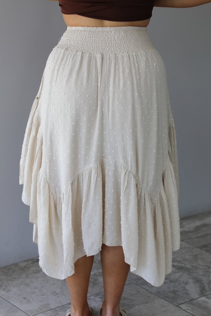 Woman wearing a sand colored midi skirt featuring a smocked waistline, a polka dot textured material, a studded detail, and a midi length. The photo is taken indoors against a white wall with white marble flooring and shows the back of the skirt. 