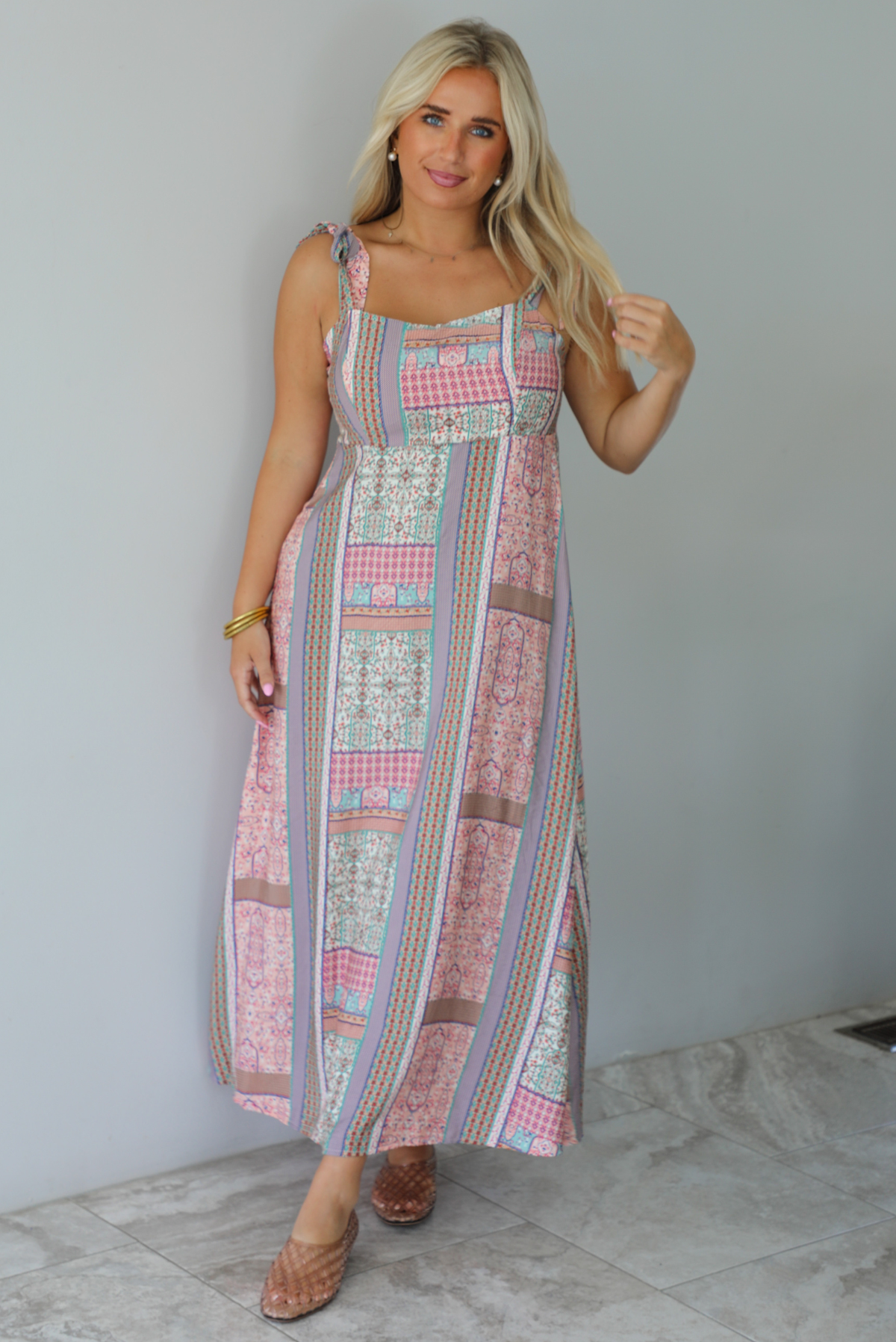 A woman wearing a thick strapped maxi dress with a patchwork print of pink gingham, beige paisley, and blue patterns, standing indoors against a white wall with white marble flooring. 