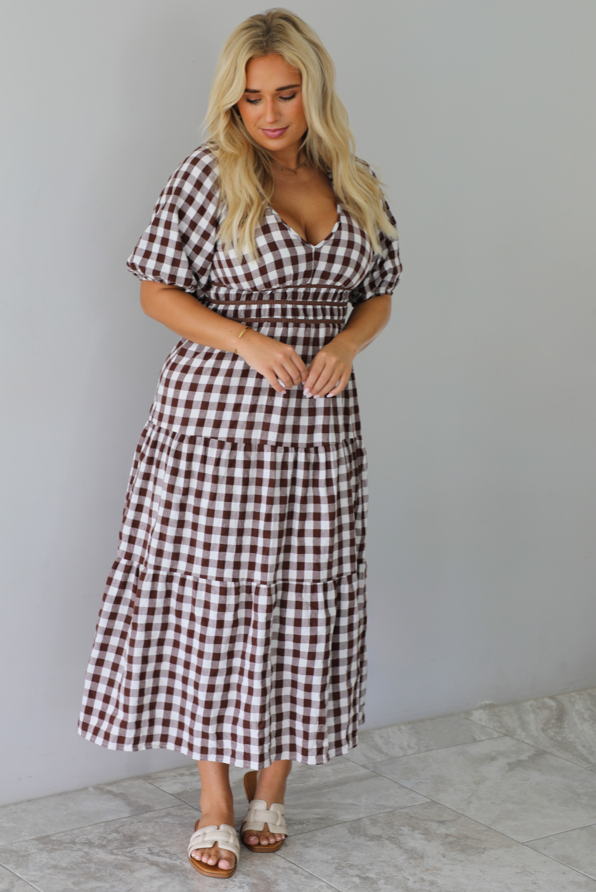 A woman modeling a brown and white gingham tiered midi dress with puff sleeves and a smocked waist, paired with beige slide sandals, standing indoors against a white wall with white marble flooring. 