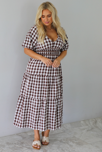 A woman modeling a brown and white gingham tiered midi dress with puff sleeves and a smocked waist, paired with beige slide sandals, standing indoors against a white wall with white marble flooring. 