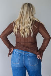A woman shown from the back wearing a sheer brown lace long sleeved top with a quarter-button closure detail and a ruffled detail at the top of the bodice, untucked to show the full length,  paired with dark wash denim with a black belt, standing indoors against a white wall.