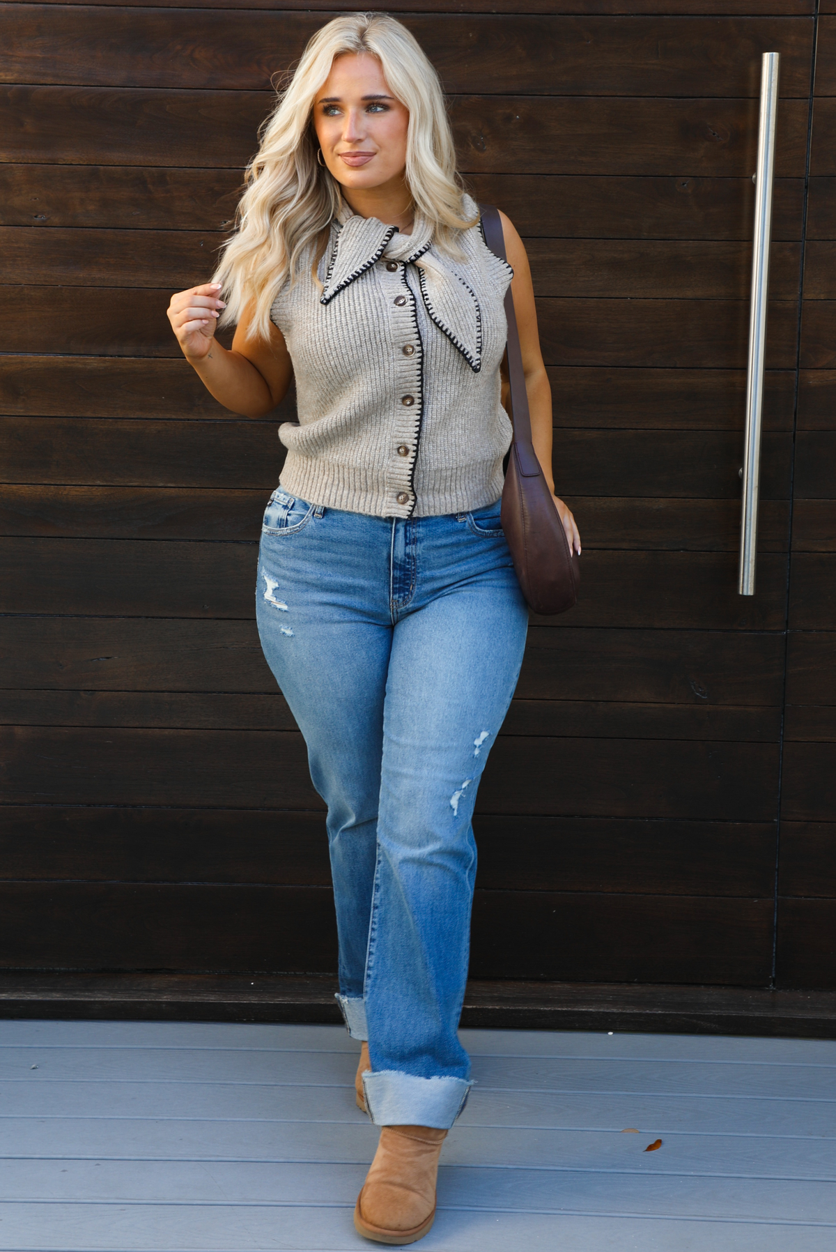 Woman model wearing a taupe sweater vest with black stitched detailing, has a tie detail at the neckline, is sleeveless, has functional button closures in the front, and has a ribbed texture. The sweater vest is paired with a medium wash denim, a dark brown leather purse, and ugg boots. The photo is taken outdoors against a dark brown wooden door. 