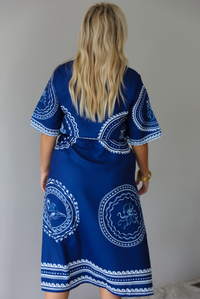 A woman shown from behind wearing a navy blue midi dress with white patterned detailing of waves and various sea creatures in circled patterns along the dress with a collared neckline, a tied waistline, and functional button closures along the front, standing indoors against a white wall with white marble flooring. 