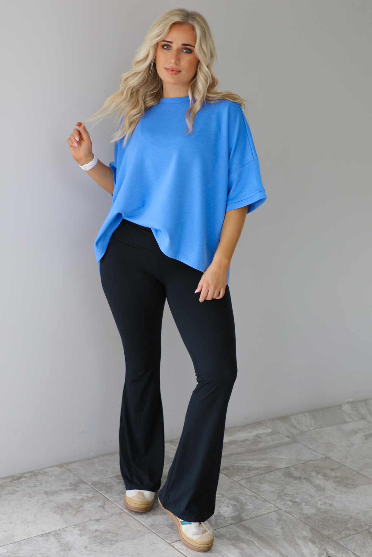 Woman wearing an oversized blue box tee with black leggings that flare slightly at the hem. The photo is taken indoors against a white wall with white marble flooring. 