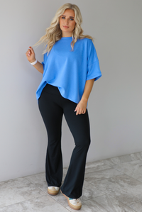 Woman wearing an oversized blue box tee with black leggings that flare slightly at the hem. The photo is taken indoors against a white wall with white marble flooring. 