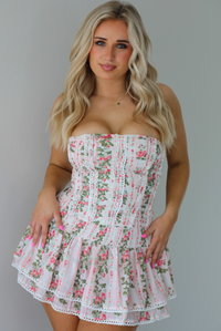 Woman wearing a white and pink floral two piece set featuring a strapless corset style top and an a ruffled skirt. The photo is taken indoors against a white wall.