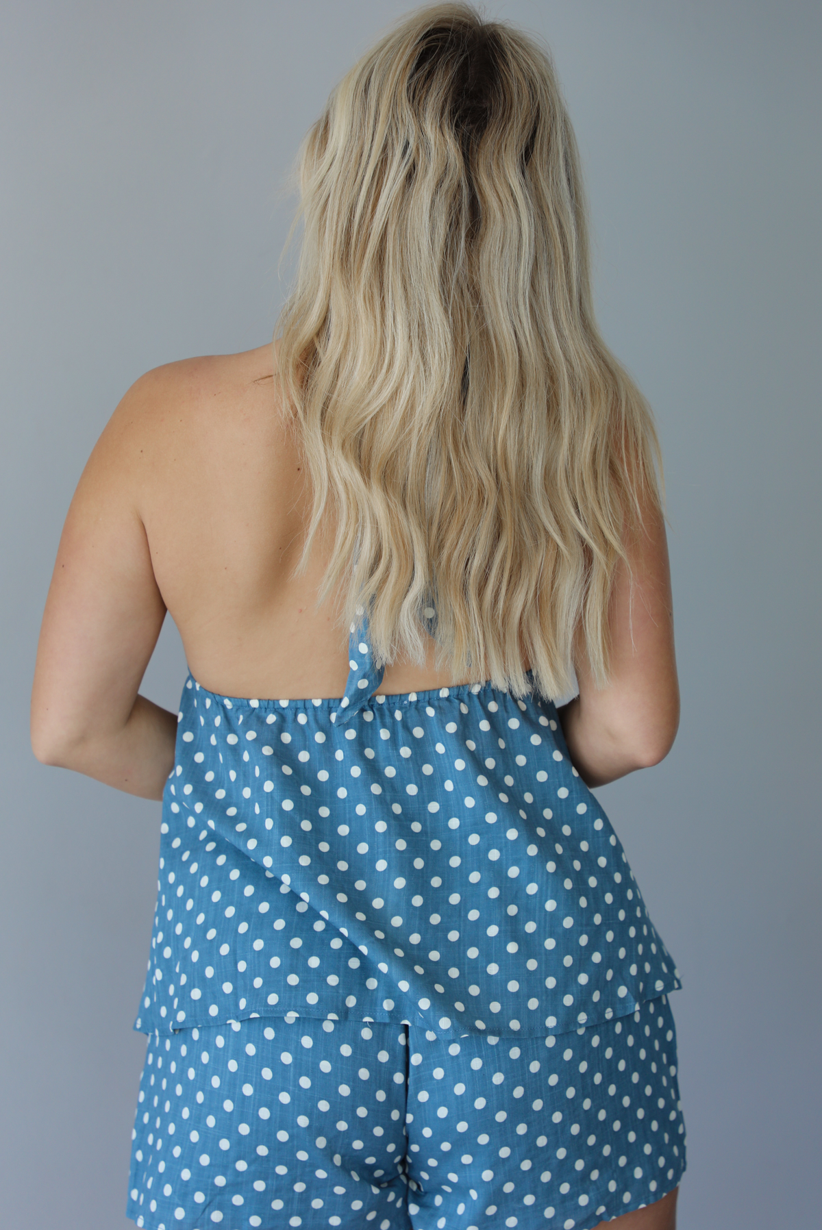 Woman wearing denim blue with white polka dot halter tie top and high waisted shorts indoors against a white wall. The photo shows the back of the top, which has an open back design and an elastic band at the waist of the top.
