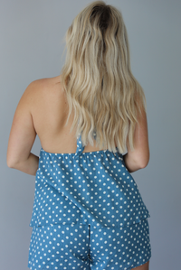 Woman wearing denim blue with white polka dot halter tie top and high waisted shorts indoors against a white wall. The photo shows the back of the top, which has an open back design and an elastic band at the waist of the top.