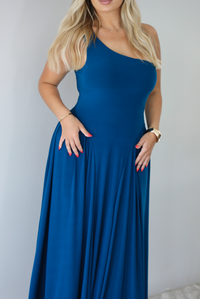 Woman wearing a blue one strap maxi dress indoors against a white wall. The photo shows a closeup of the bodice and waist of the dress. 